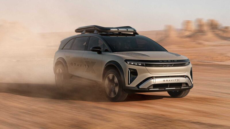 A silver Lucid Gravity SUV, inspired by its Pebble Beach debut, drives on a dusty desert terrain with a roof rack, raising dust behind it under clear skies—showcasing rugged upgrades and trail-ready spirit.