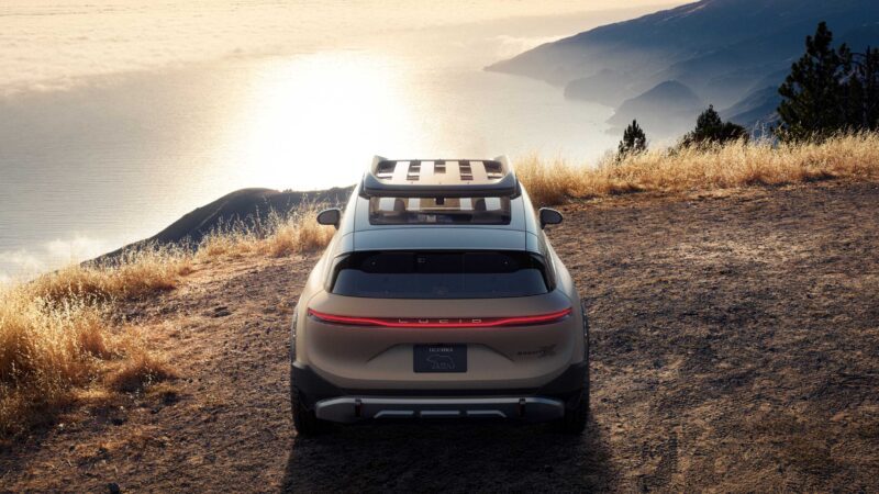 A silver SUV with a roof rack is parked on a dirt overlook above the ocean, resembling the Pebble Beach Debut: Rugged Lucid Gravity X Concept with 828 HP and trail-ready upgrades, cliffs and coastline glowing at sunset in the background.