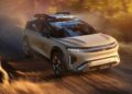 A silver Lucid Gravity X Concept with a roof cargo box drives on a dirt road through a sunlit forest, showcasing its Pebble Beach debut and trail-ready upgrades with 828 HP.