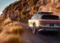 A white SUV with a roof box, inspired by the Pebble Beach Debut: Rugged Lucid Gravity X Concept, drives along a winding dirt road beside a rocky hillside under a clear sky.
