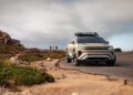 A modern SUV with a roof rack is parked on a coastal road, channeling the Pebble Beach debut of the rugged Lucid Gravity X Concept, breaking cover with 828 HP and trail-ready upgrades against rocky terrain and cloudy skies.