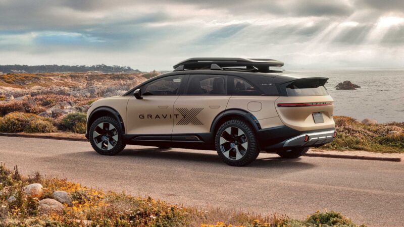 A beige electric SUV with "GRAVIT" branding is parked on a coastal road, evoking the Pebble Beach Debut: Rugged Lucid Gravity X Concept Breaks Cover With 828 HP & Trail-Ready Upgrades—rocky cliffs and ocean under a cloudy sky set the scene.