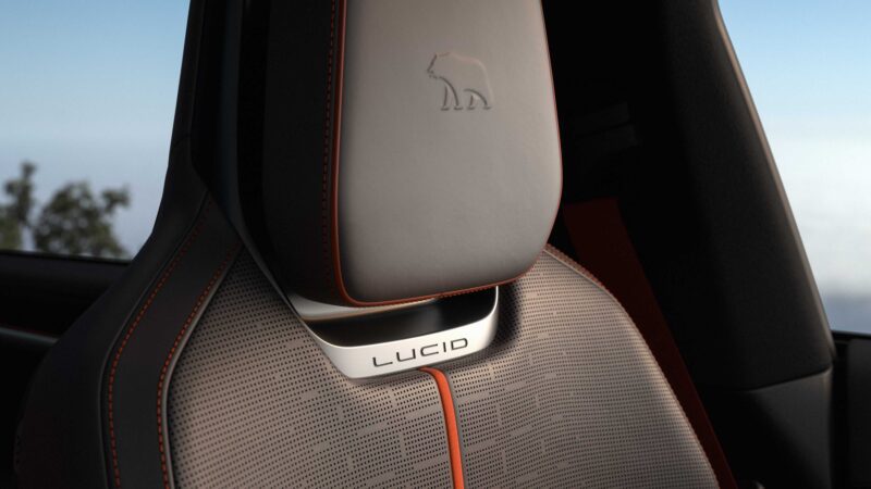 Close-up of a car seat with grey upholstery, orange stitching, a bear logo on the headrest, and "LUCID" branding on the seat back—features showcased in the Pebble Beach Debut: Rugged Lucid Gravity X Concept with 828 HP & trail-ready upgrades.