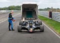 A man in racing gear stands beside a vintage black Lotus Type 79 John Player Special Formula 1 car parked in front of an open trailer on a racetrack.