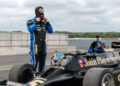 A person in a racing suit adjusts their balaclava beside a vintage black and gold Lotus Type 79 "John Player Special" Formula 1 car on a racetrack, reminiscent of Jan Lammers’ era.