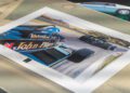 A photograph of a Formula 1 race car print featuring the iconic Lotus Type 79 John Player Special on track, placed on a table with another car image—possibly featuring Jan Lammers—partly visible in the background.
