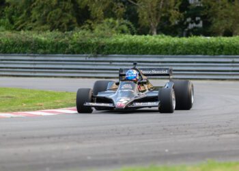 A black Lotus Type 79 Formula 1 car with John Player Special livery takes a corner on a racetrack, driven by Jan Lammers wearing a blue helmet.