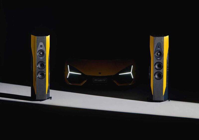 Two black and yellow speakers, part of the Only 50 Worldwide: Lamborghini x Sonus Faber Il Cremonese Ex3me series, stand in front of a yellow Lamborghini with illuminated headlights against a dark background.