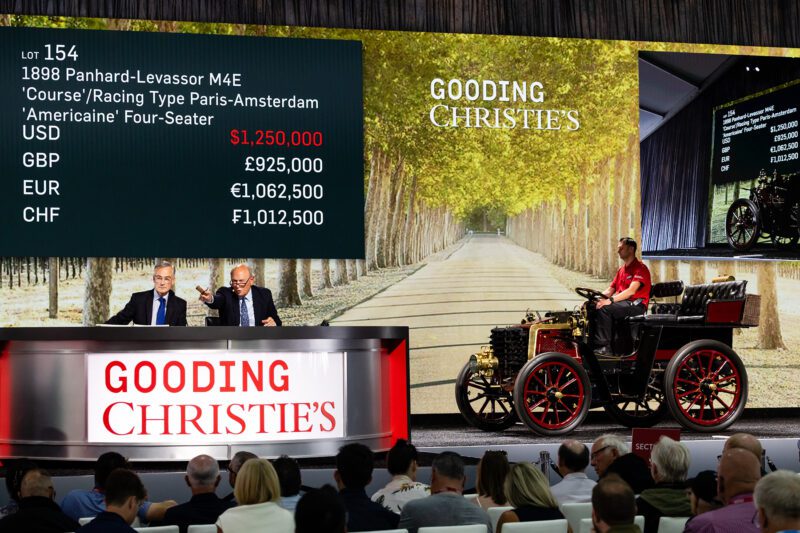 Two auctioneers sit at a desk as a vintage 1898 Panhard-Levassor is auctioned at Gooding Christie’s Pebble Beach Auction, with bids displayed on a screen in multiple currencies.
