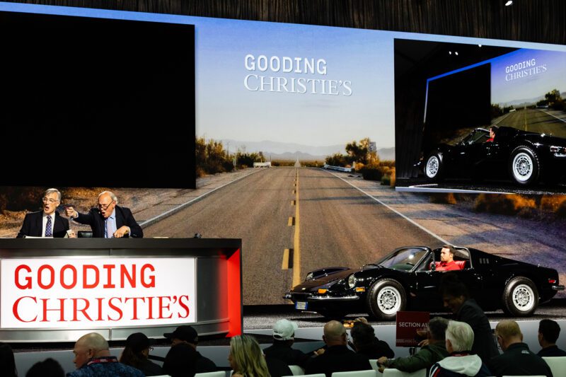 An auction scene at Gooding & Company’s Pebble Beach Auction, with two auctioneers at a desk and a sleek black Ferrari 250 GT California Spider, driven on stage and highlighted on a large screen.