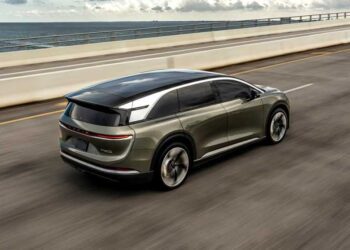 A green electric SUV with impressive autonomy drives on a highway next to the ocean under a partly cloudy sky.