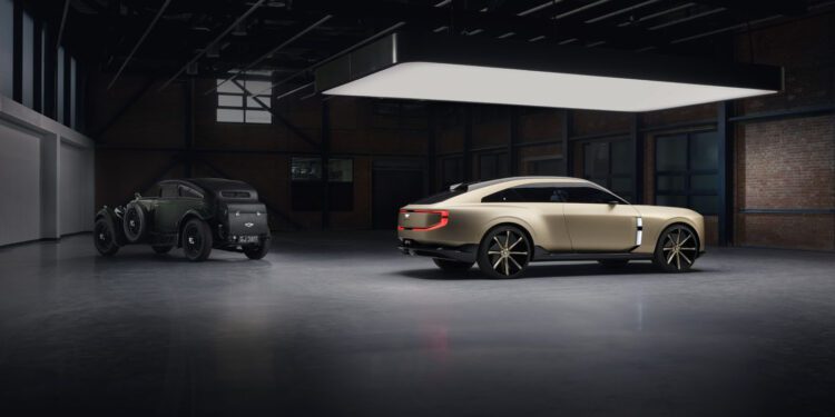 A vintage black car and a modern beige Bentley EXP 15 Concept are parked side by side in a spacious, dimly lit industrial garage, capturing the spirit of Monterey Car Week.