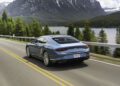 A blue Bentley coupe, inspired by the EXP 15 Concept, drives on a highway beside a lake and forested mountains under a partly cloudy sky.