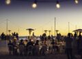 People are dining at tables on a beach at sunset, with string lights overhead and heaters placed around, creating a festive outdoor atmosphere reminiscent of Monterey Car Week elegance.