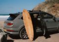 A wooden surfboard is propped against a grey Bentley SUV with an open door, parked near a rocky beach with waves in the background—an elegant scene that wouldn’t look out of place at Monterey Car Week.