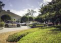 Several luxury cars, including a Bentley EXP 15 Concept, are parked outside a modern house as guests mingle and converse in the sunlit, landscaped area during Monterey Car Week.