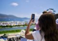 A person holding up a smartphone to take a photo at an outdoor event near water, with blurred crowds and mountains in the background during Monterey Car Week.