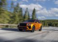 An orange Bentley EXP 15 Concept luxury SUV drives on an empty highway surrounded by greenery and trees under a partly cloudy sky, capturing the spirit of Monterey Car Week.
