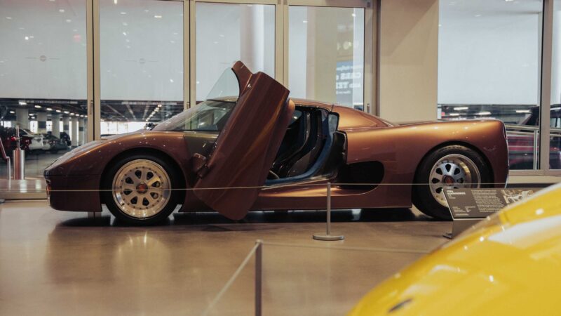 A brown sports car with its driver's door open is displayed indoors behind a rope barrier at the Petersen Automotive Museum, next to a yellow car, with glass windows in the background.