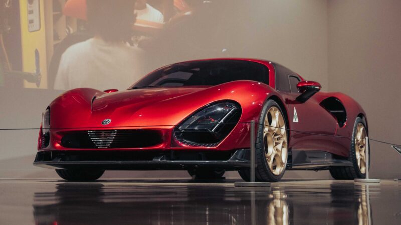 A sleek red sports car with gold wheels is displayed indoors on a polished floor at the Petersen Automotive Museum, with a blurred projection in the background.
