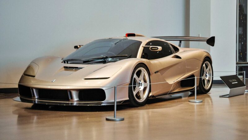 A silver sports car, one of the headliners at the Petersen Automotive Museum, is displayed indoors behind stanchions, with a sign and large windows visible in the background.