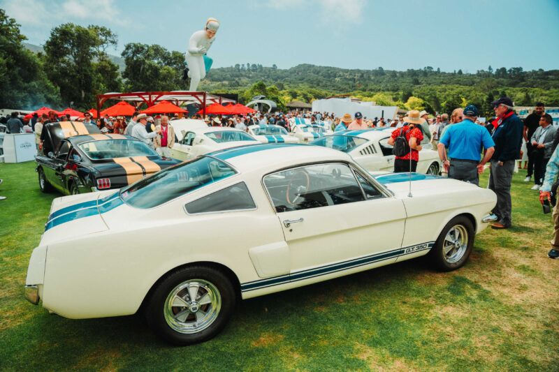 A classic white Ford Mustang GT350 with blue stripes is displayed on a grassy lawn at a crowded outdoor car show, where the Ferrari F50 GT1 was crowned Best of Show at The Quail A Motorsports Gathering 2025.