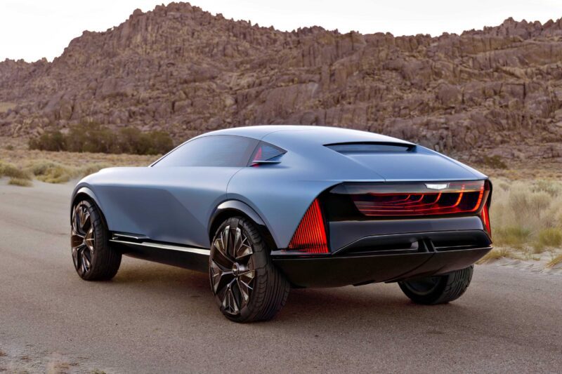 A futuristic matte blue SUV concept car, inspired by Cadillac Elevated Velocity luxury EVs, is parked on a desert road with rocky hills in the background.