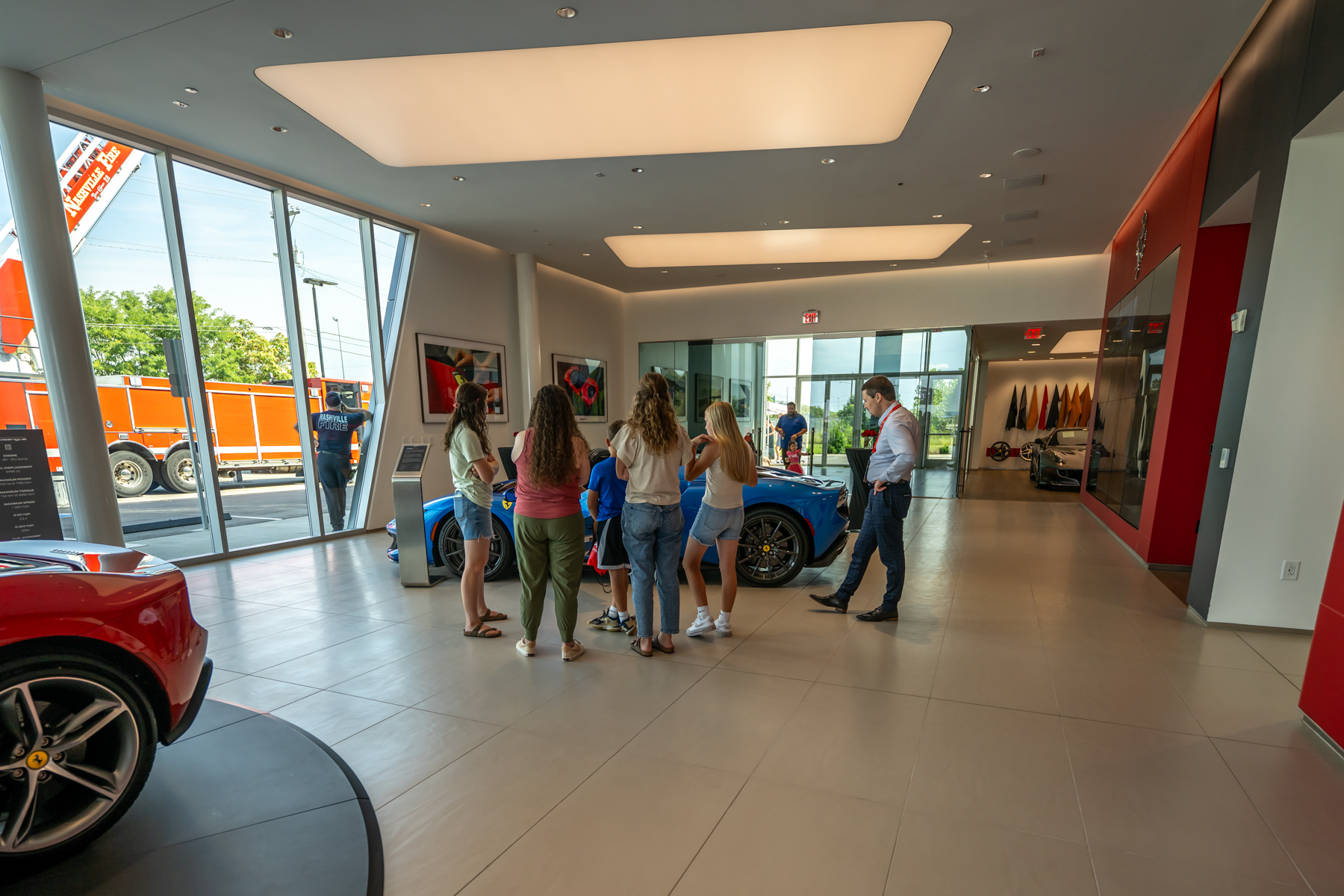 A group of people stands near a blue sports car inside a modern car showroom with large windows, where dreams and Wishes in Motion come alive among the cars on display.
