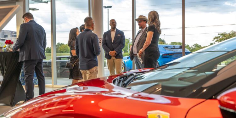 A group of people stand and converse in a car showroom next to a red Ferrari, their wishes in motion as evening light streams through large windows showing cars and trees outside.