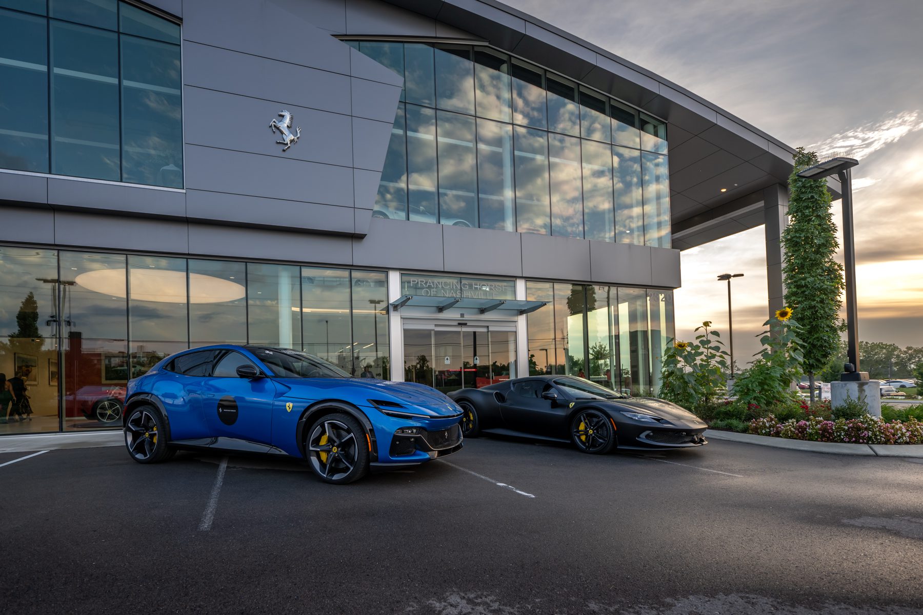 A blue Ferrari SUV and a black Ferrari sports car stand parked in front of a modern glass Ferrari dealership at sunset, turning evening wishes in motion into unforgettable dreams.