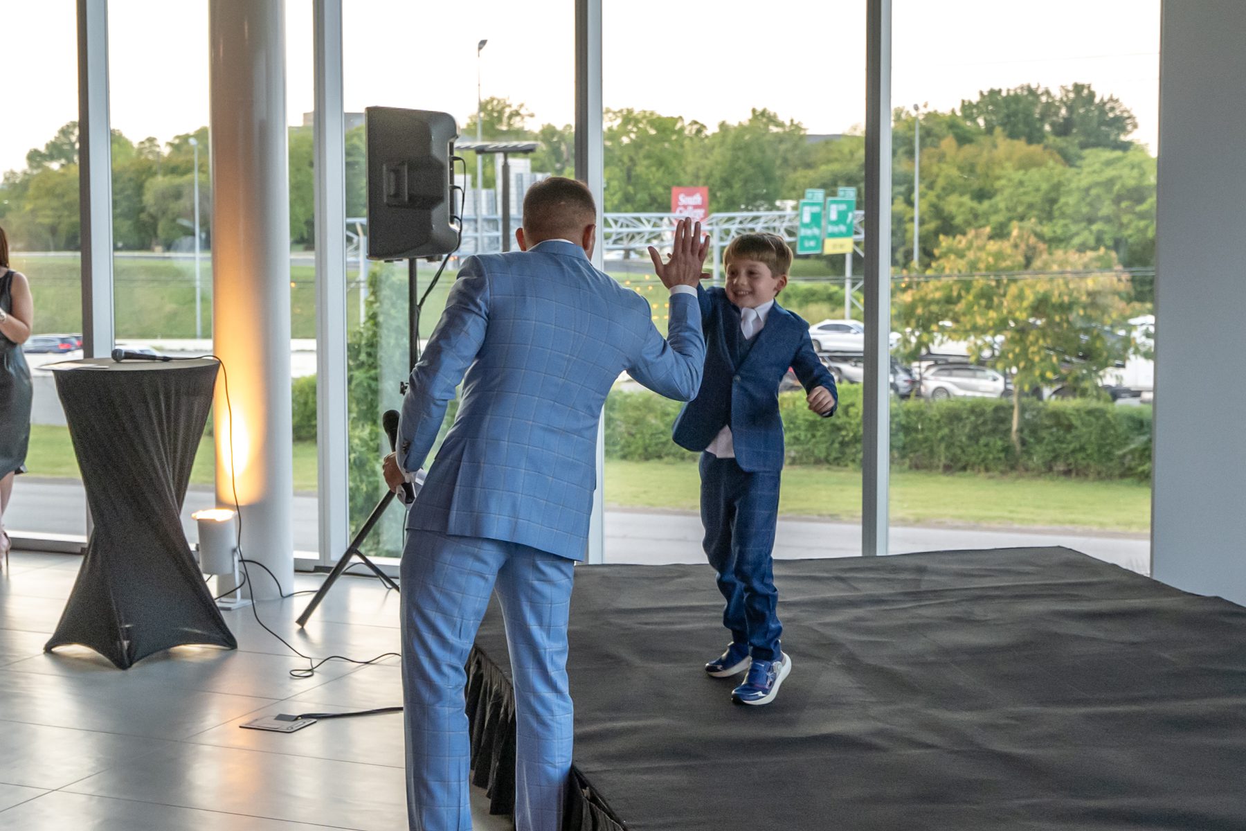 A man and a boy in blue suits exchange a high five on stage, framed by large windows overlooking a parking lot and greenery, as Dreams come alive in this inspiring moment.