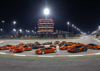 A group of orange sports cars, part of The Most Extraordinary McLaren Collection On The Planet Just Sold To A Single Buyer, is parked on a racetrack at night with an illuminated tower in the background. A person stands in front of the cars.