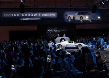 A white and blue sports car is displayed on stage at the Broad Arrow Jet Center Auction: Top 10 Results And Highlights event, surrounded by a seated audience and large automotive banners.