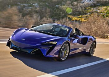 A blue convertible sports car, like the 2025 McLaren Artura Spider, speeds along a road with blurred trees and rocky terrain in the background—perfectly capturing the droptop’s split personality in motion.