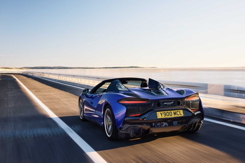 A 2025 McLaren Artura Spider cruises as a blue convertible on an empty road near the water under a clear sky, embodying the droptop with a split personality described in recent reviews.