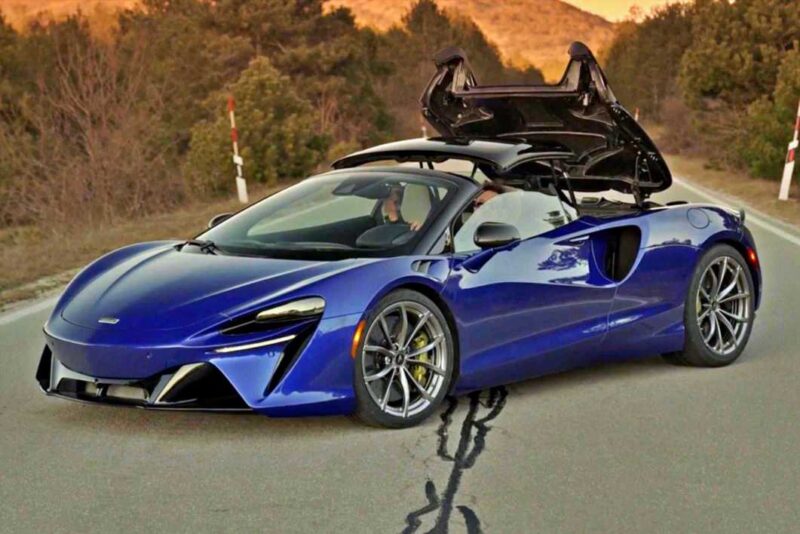 A blue 2025 McLaren Artura Spider with its driver-side butterfly door open is parked on a road, surrounded by trees and mountains—a perfect setting for a review of this droptop with a split personality.