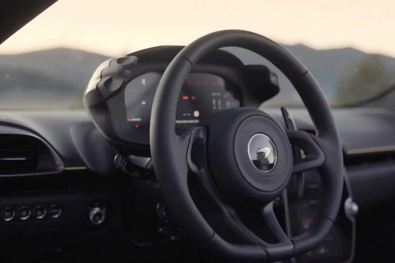 Close-up view of a luxury car’s steering wheel and digital dashboard, with mountains visible through the windshield—perfectly capturing the essence of the 2025 McLaren Artura Spider Review: Droptop With A Split Personality.