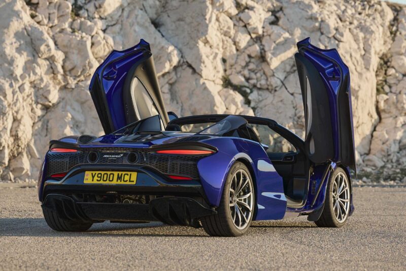 A blue 2025 McLaren Artura Spider, its scissor doors open wide, is parked in front of a rocky cliff on a sunny day—a true droptop with a split personality.