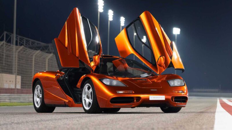 An extraordinary McLaren with both butterfly doors open is parked on a racetrack at night under bright lights.