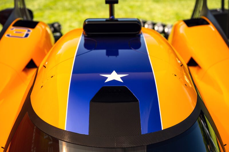 Close-up of an orange race car with a central blue stripe featuring a white star, viewed from above, as McLaren’s Project: Endurance Hypercar debuts its Triple Crown livery at Monterey, with a grassy area in the background.