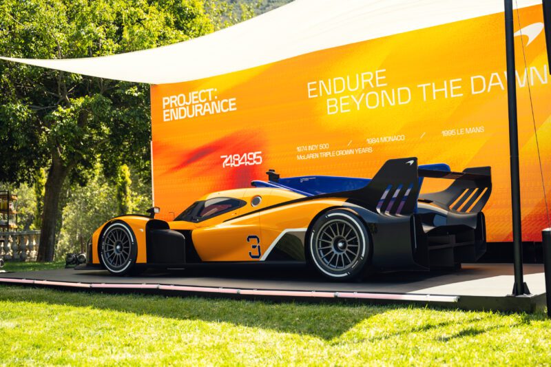 A sleek orange and black race car, McLaren’s Project: Endurance Hypercar, debuts its Triple Crown livery on a platform in front of an orange "Project: Endurance" billboard reading "Endure Beyond the Dawn.