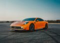 An orange Mercedes-AMG GT sports car is parked on pavement under a clear blue sky, with greenery and landscape in the background—reminiscent of the legendary 24-Hour Distance World Record set at Nardò.