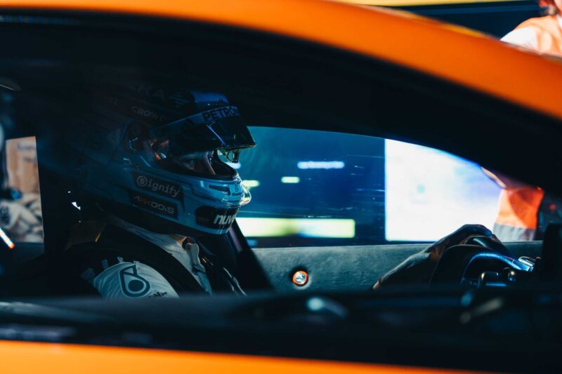 A race car driver wearing a helmet sits in the driver's seat of an orange Mercedes-AMG GT at Nardò, focused on the road ahead, with hands on the steering wheel, ready to chase the 24-Hour Distance World Record.