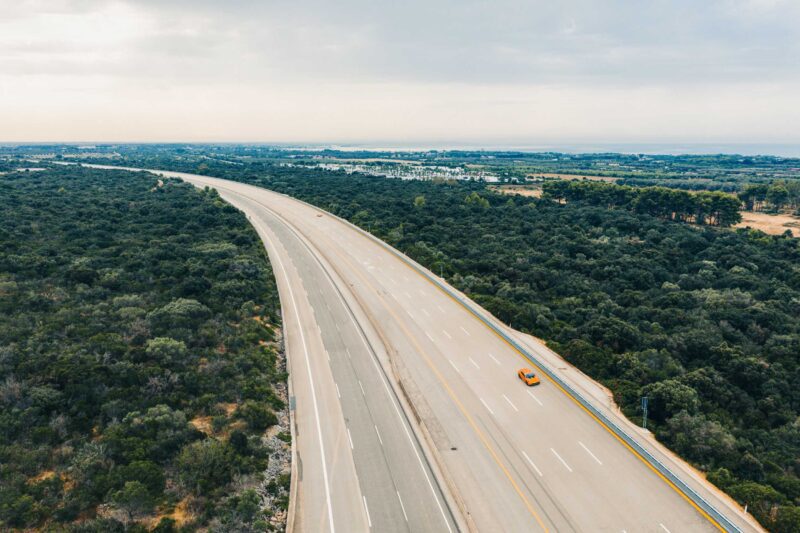 Aerial view of an empty multi-lane highway at Nardò, surrounded by dense greenery, with a single yellow Mercedes-AMG GT driving along one lane under a cloudy sky—an ideal scene for a 24-Hour Distance World Record attempt.
