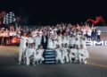 A large group, some in white racing suits, pose on the Nardò racetrack at night with a checkered flag and AMG signs, celebrating the 24-Hour Distance World Record with the Mercedes-AMG GT as red flares light up the crowd.