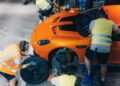 Four people in safety vests change the front wheel of an orange Mercedes-AMG GT in a workshop, echoing the energy of a 24-Hour Distance World Record attempt at Nardò.