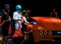 A race car driver in a white suit stands beside several crew members working on an orange Mercedes-AMG GT at Nardò, with one person leaning into the driver's side door, preparing for a 24-Hour Distance World Record attempt.