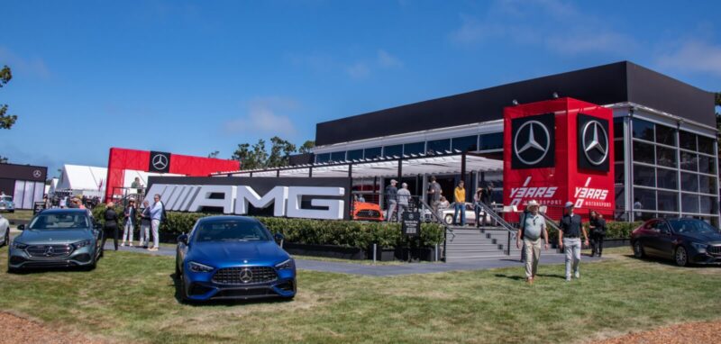 People walk around a Mercedes-AMG outdoor exhibit at Monterey Car Week 2025, featuring several cars, large AMG signage, and a building with Mercedes-Benz logos on a sunny day.