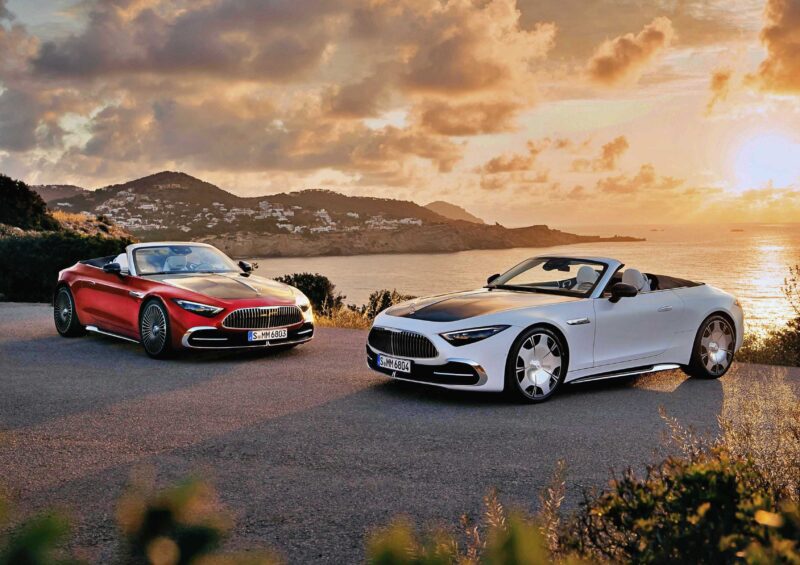 Two ultra-luxury roadsters, including the 2026 Mercedes-Maybach SL 680, are parked on a scenic coastal road at sunset, with hills and a shimmering body of water in the background.