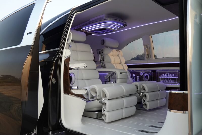 The image shows the luxurious interior of a Mercedes-Benz Vision V concept van, featuring white leather reclining seats, wood accents, built-in screens, and an illuminated minibar.
